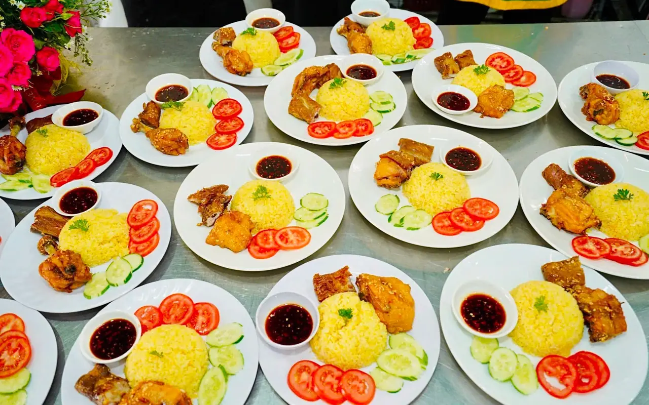 Nhiều đĩa cơm gà xối mỡ bày trên bàn bếp inox: gà chiên vàng giòn, cơm chiên vàng, cà chua, dưa leo và chén nước chấm — Bếp Cô Linh