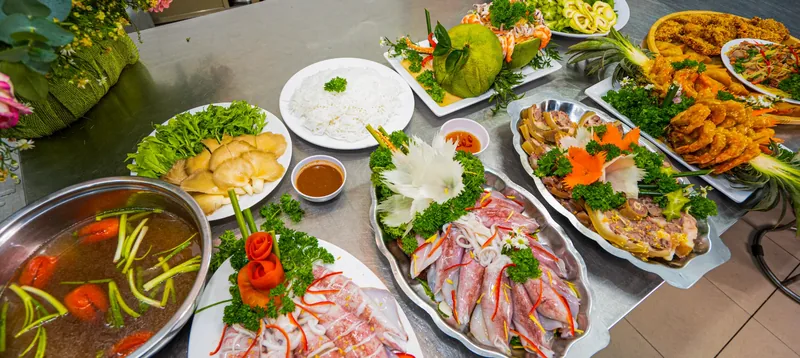 Các món tiệc bày trên bàn bếp: nồi lẩu nước chua, mực hấp, giò, tôm chiên, đĩa rau nấm và bún