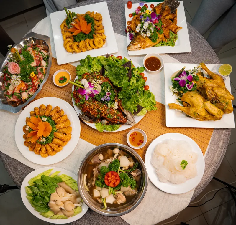 Bàn tròn bày đủ món tiệc: mực chiên, cá chiên, gà, chả giò, một món cuốn, lẩu cùng rau nấm và bún