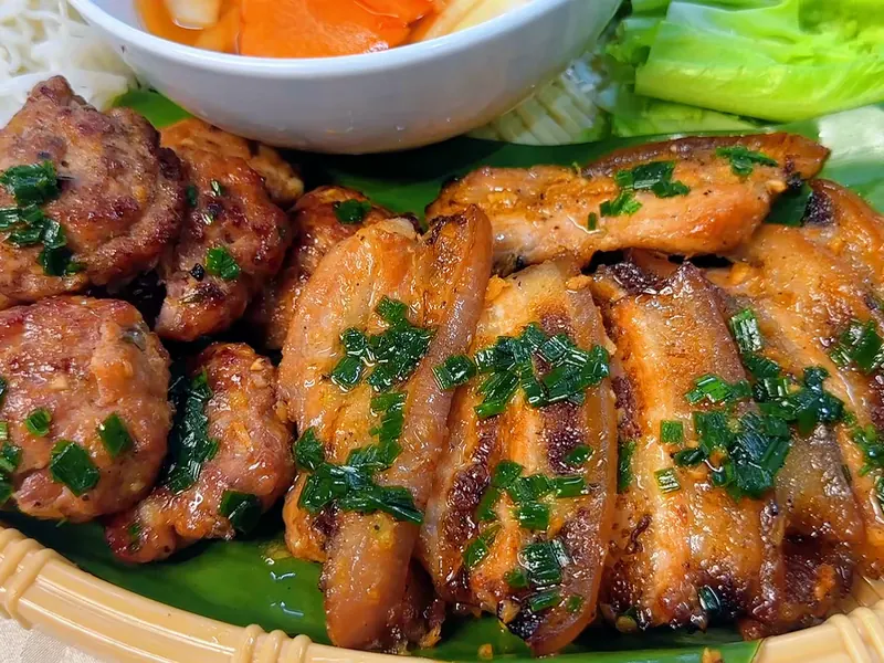 Cận cảnh chả viên và chả miếng ba rọi nướng vàng rắc hành lá trên mẹt lót lá chuối — khung hình từ video khoá Bún Chả Hà Nội