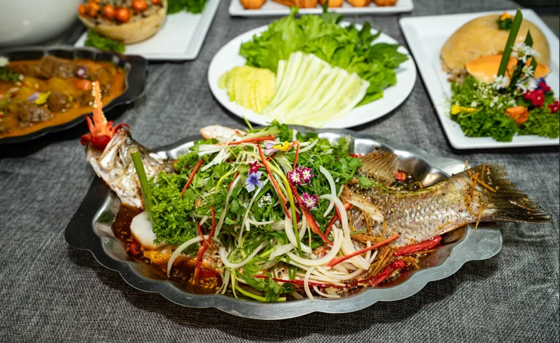 Cá chẽm hấp Triều Châu phủ hành, rau thơm và hoa ăn được, bày trên khay inox