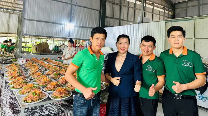 Cô Linh đứng cùng ba học viên nam trong đồng phục xanh cam, phía sau là bàn dài bày sẵn hàng chục đĩa gỏi trang trí hoa cà rốt