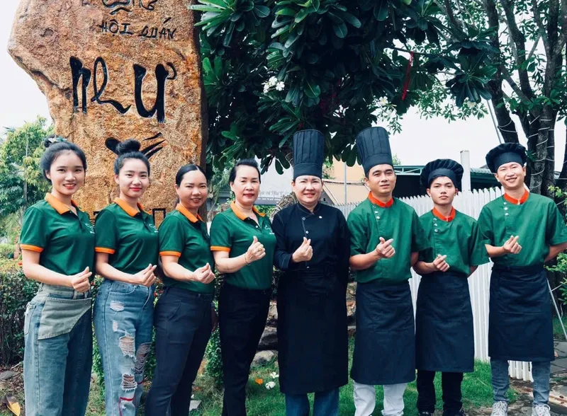 Cô Linh mặc áo bếp đen đứng cùng đội bếp và phục vụ mặc đồng phục xanh lá viền cam trước một buổi tiệc