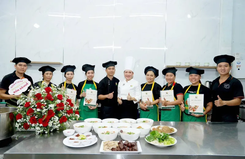 Cô Linh cùng học viên và đội ngũ chụp ảnh lưu niệm sau buổi học bún bò Huế, cầm sổ công thức, phía trước là các tô bún thành phẩm