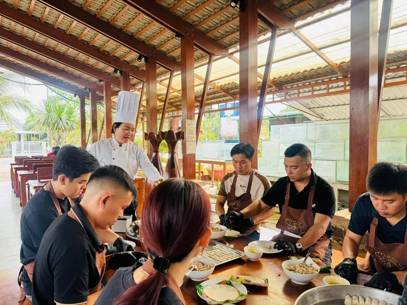 Cô Linh mặc áo bếp trắng hướng dẫn các học viên cuốn chả giò tại lớp