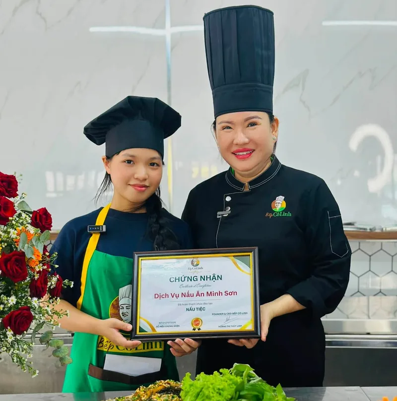 Cô Linh trao chứng nhận hoàn thành khoá đào tạo nấu tiệc cho một học viên còn nhỏ tuổi, phía trước là các món tiệc bạn đã nấu