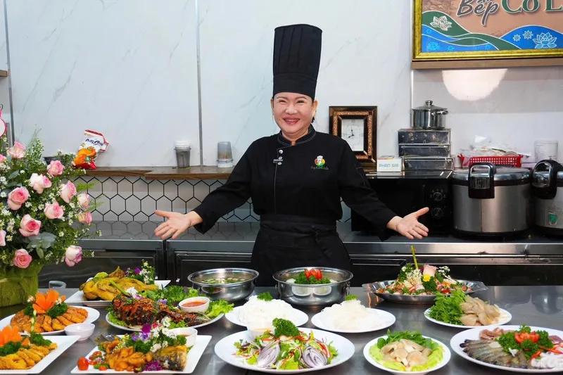 Cô Linh mặc đồng phục bếp đen, dang tay trước bàn bày nhiều món tiệc như gà, cá chiên, gỏi, súp và hải sản tại bếp Bếp Cô Linh