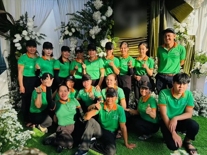 Đội ngũ Bếp Cô Linh mặc áo xanh cam, đội mũ đen, chụp ảnh chung trước khu trang trí hoa ở một tiệc (chữ lồng của chủ tiệc đã làm mờ)