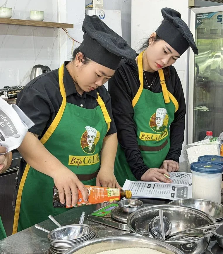 Hai học viên đeo tạp dề Bếp Cô Linh tự tay cân gia vị trên cân điện tử theo tờ công thức trong lớp học nấu tiệc