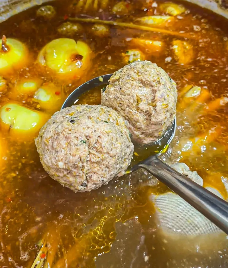 Nồi nước lèo bún bò Huế màu cam nâu, có hành củ, sả và hai viên chả lớn đang nổi