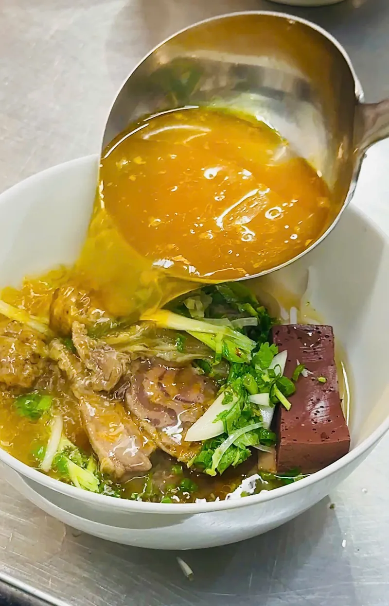 Múc nước lèo bún bò Huế vàng cam trong, rưới vào tô có thịt, huyết và hành lá