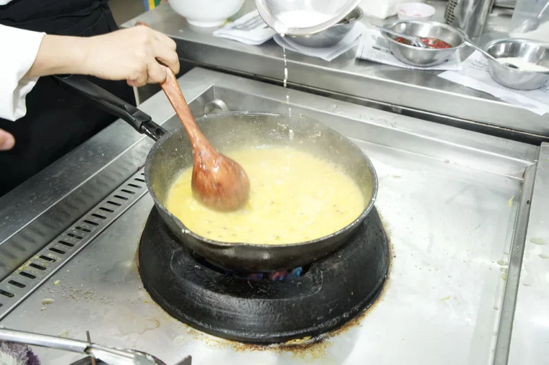 Chảo sốt vàng đang nấu trên bếp, tay cô Linh khuấy bằng muỗng trong lớp học nấu tiệc