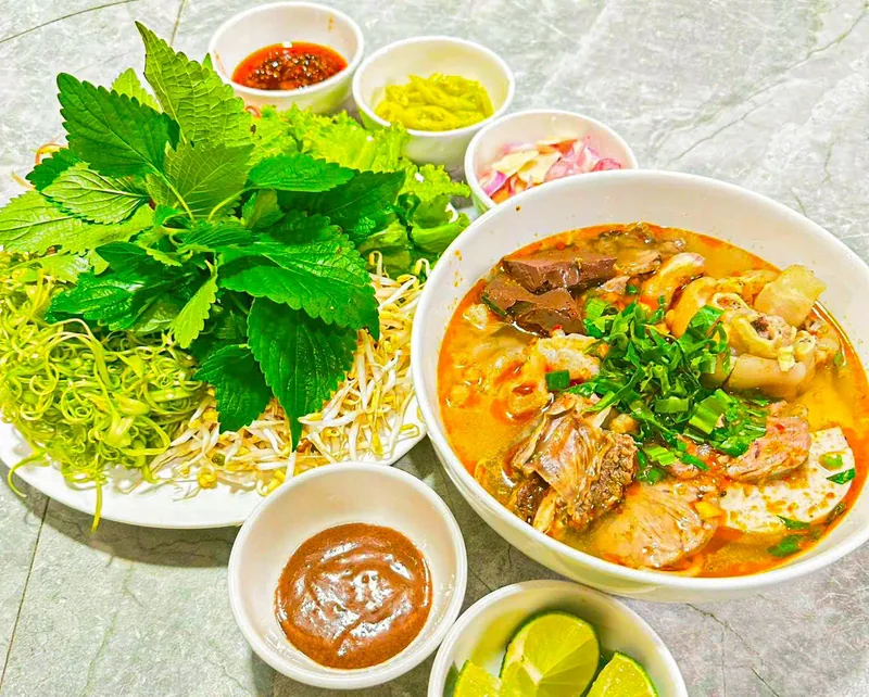 Tô bún bò Huế có giò heo, huyết, thịt và chả, ăn kèm đĩa rau sống, chén mắm ruốc, sa tế, ớt ngâm, hành tây ngâm và chanh